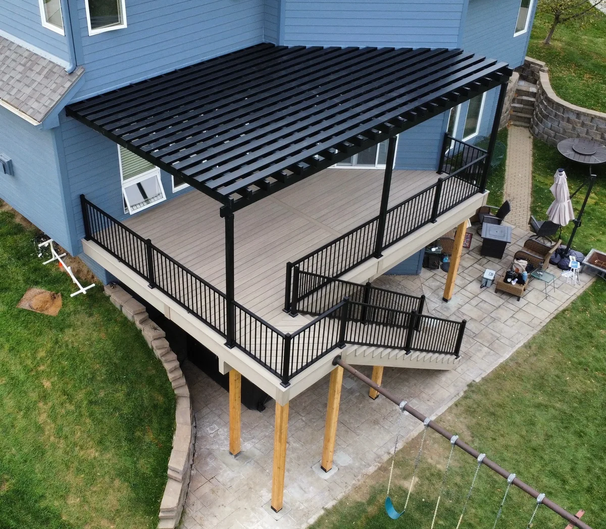 Elevated view of a deck with a black pergola, matching railings, and stairs beside a blue house